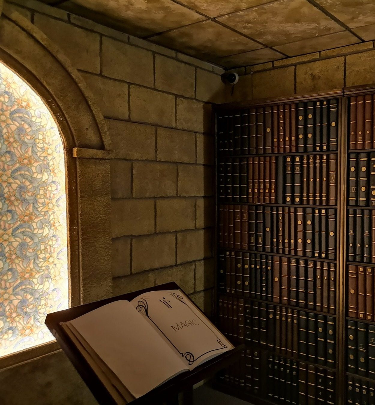 Salle L'École des Sorciers : bibliothèque de pierre, grimoire ouvert devant une fenêtre à vitraux